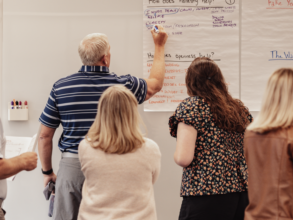 a group of three people staring at a whiteboard. one man is writing with his right hand