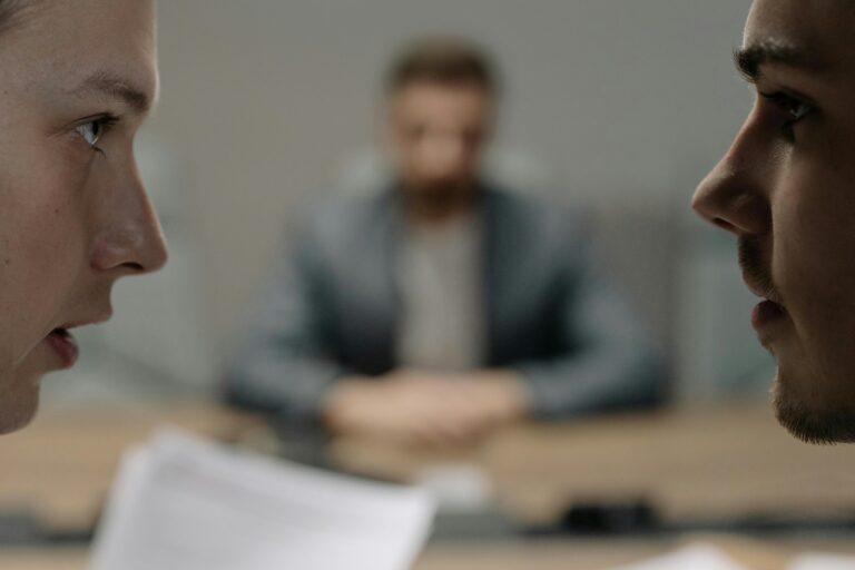 Two people facing each other in a tense workplace conversation with a third person seated in the background